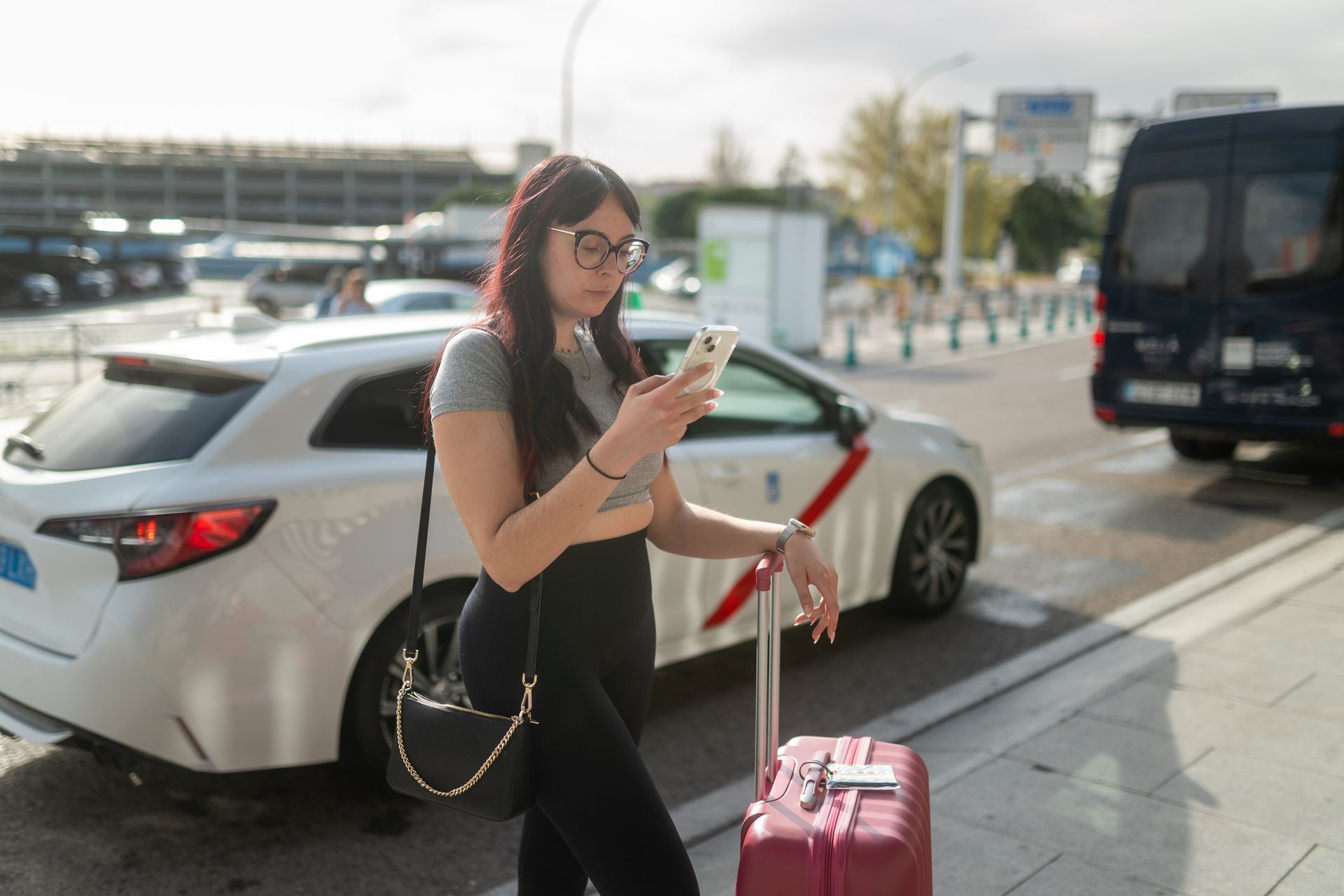 A brown-haired woman with glasses stands next to a suitcase as she checks her phone.