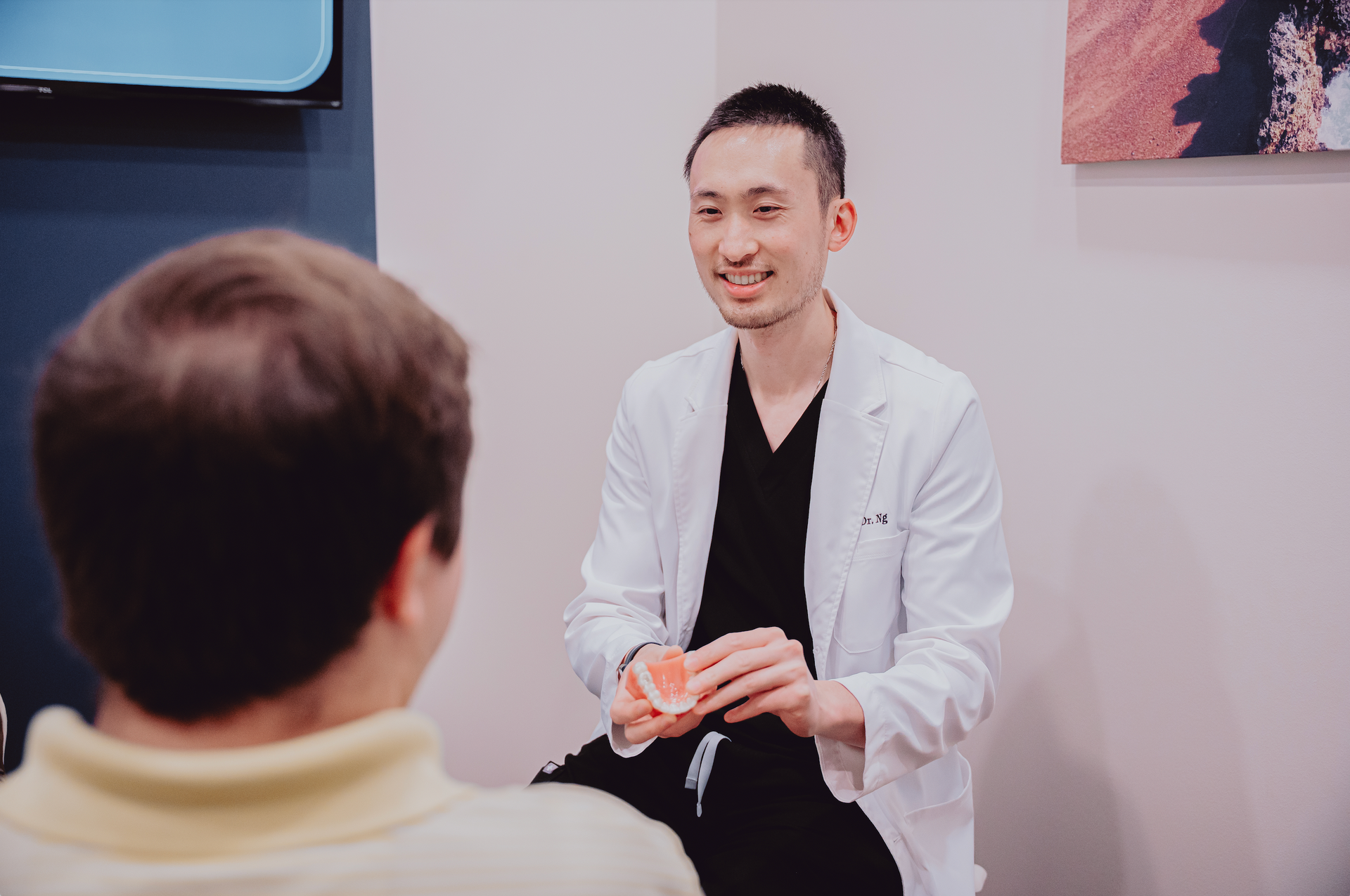 A woman is sitting in a dental chair while a dentist holds a clear retainer in his hand.