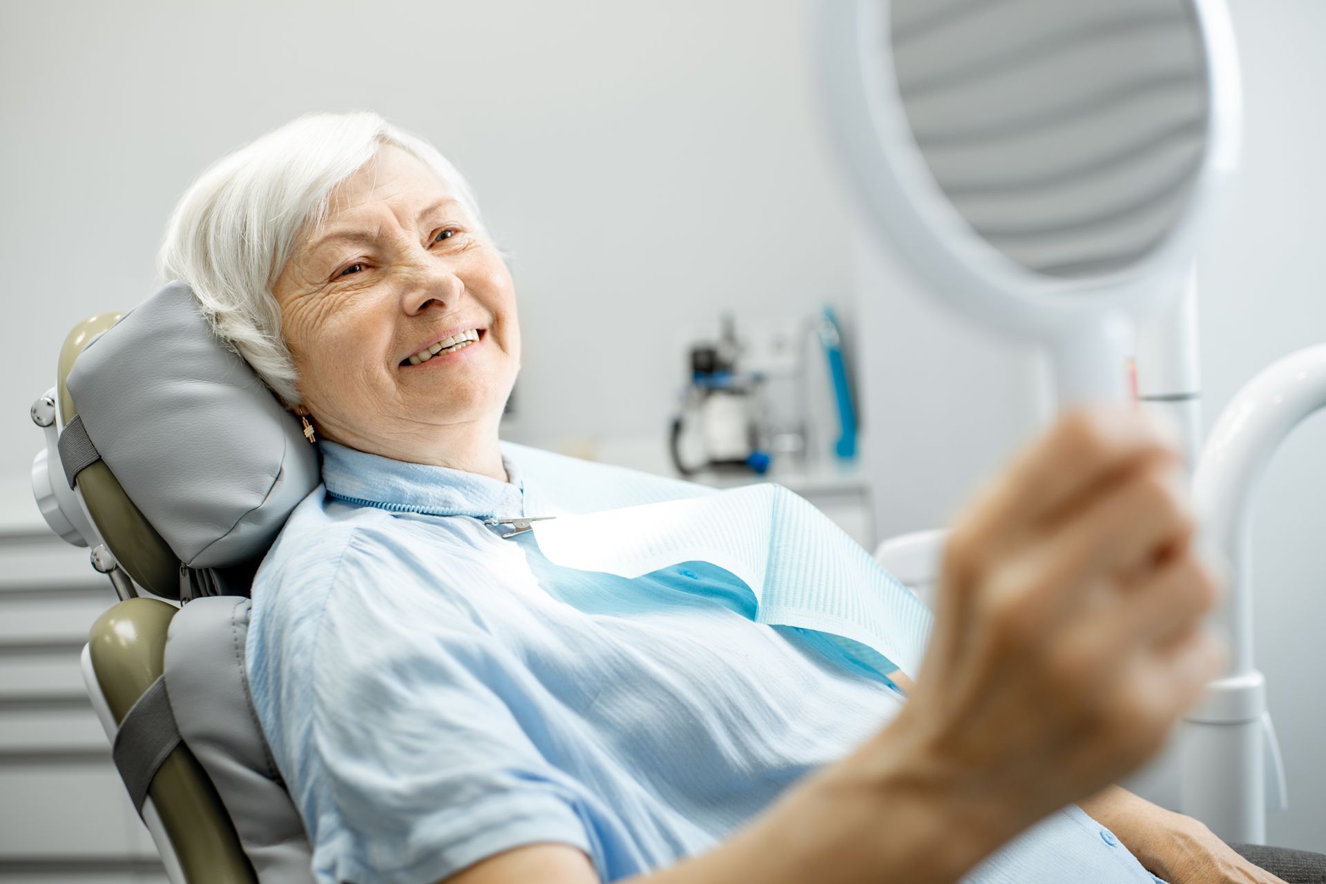 An elderly woman is sitting in a dental chair looking at her teeth in a mirror.