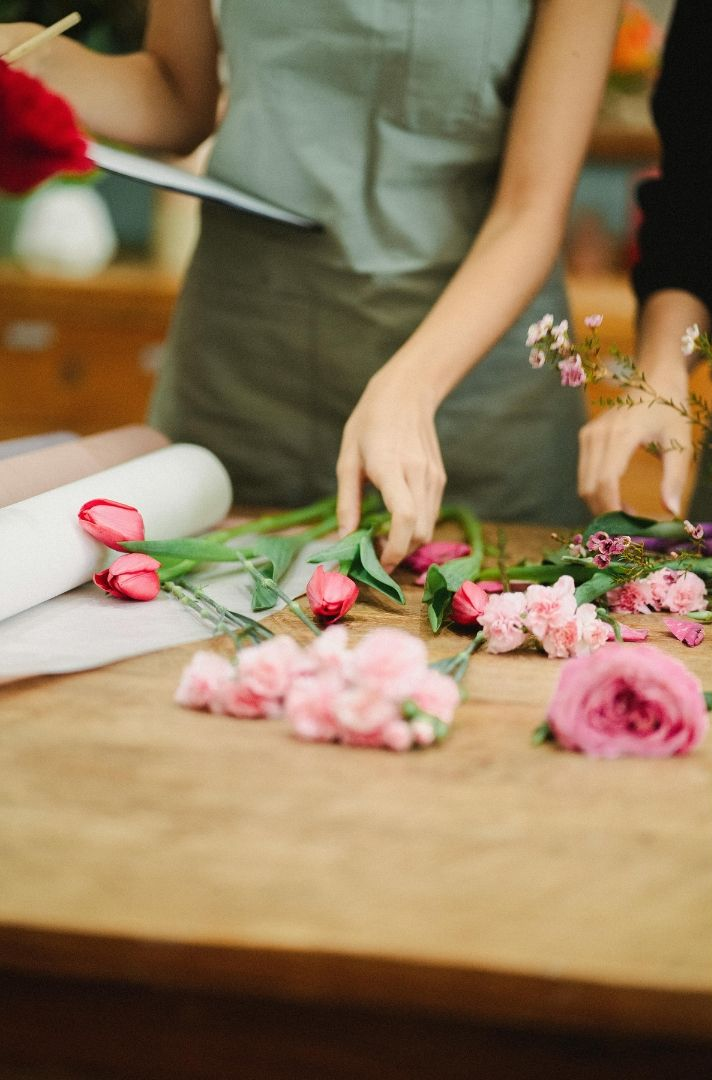 Florist arranging pink and red flowers on a wooden table, with one person wearing an apron.