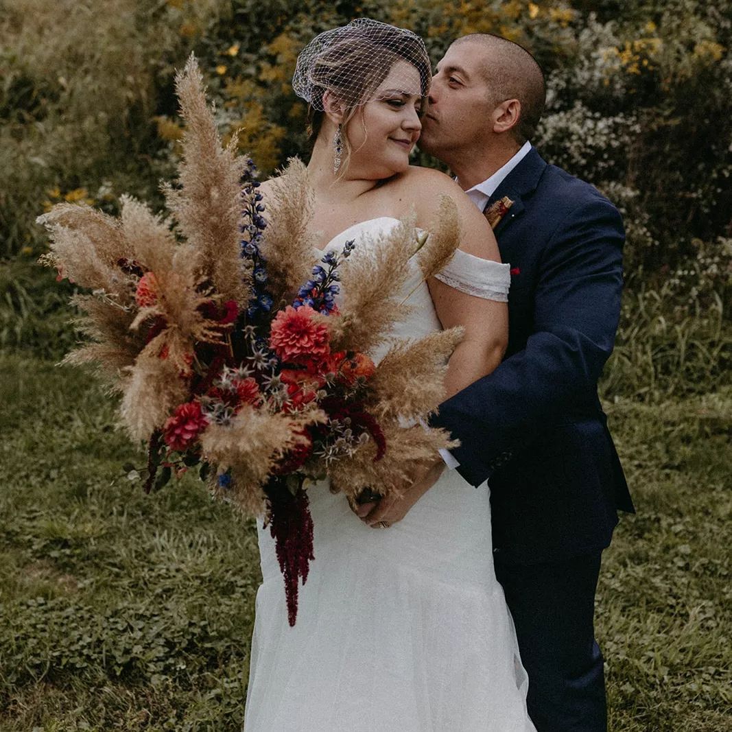 A bride and groom are kissing in a field while the bride is holding a bouquet of flowers.