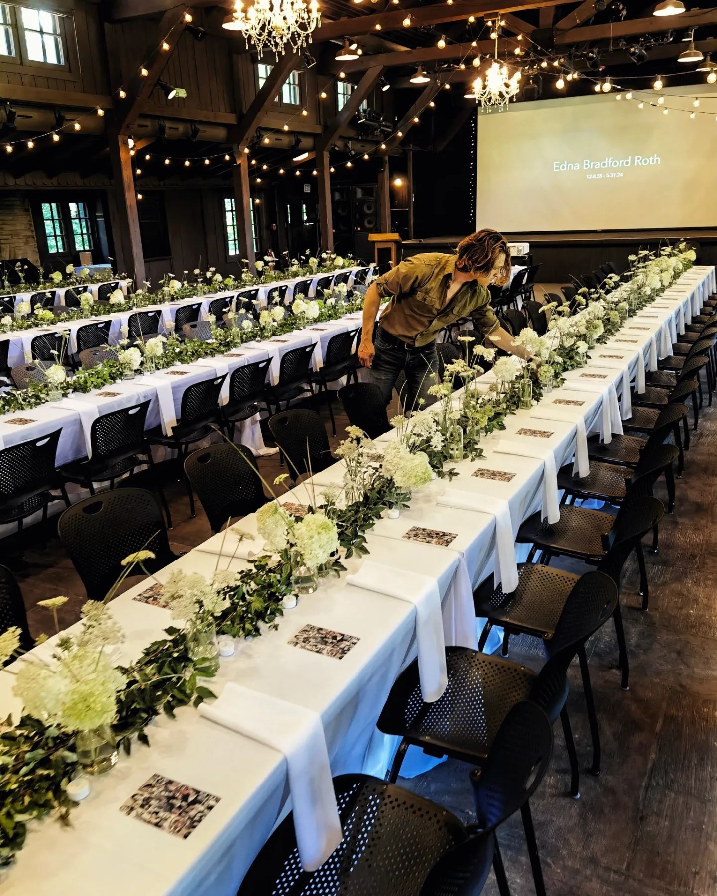 A man is standing in front of a long table with flowers on it.