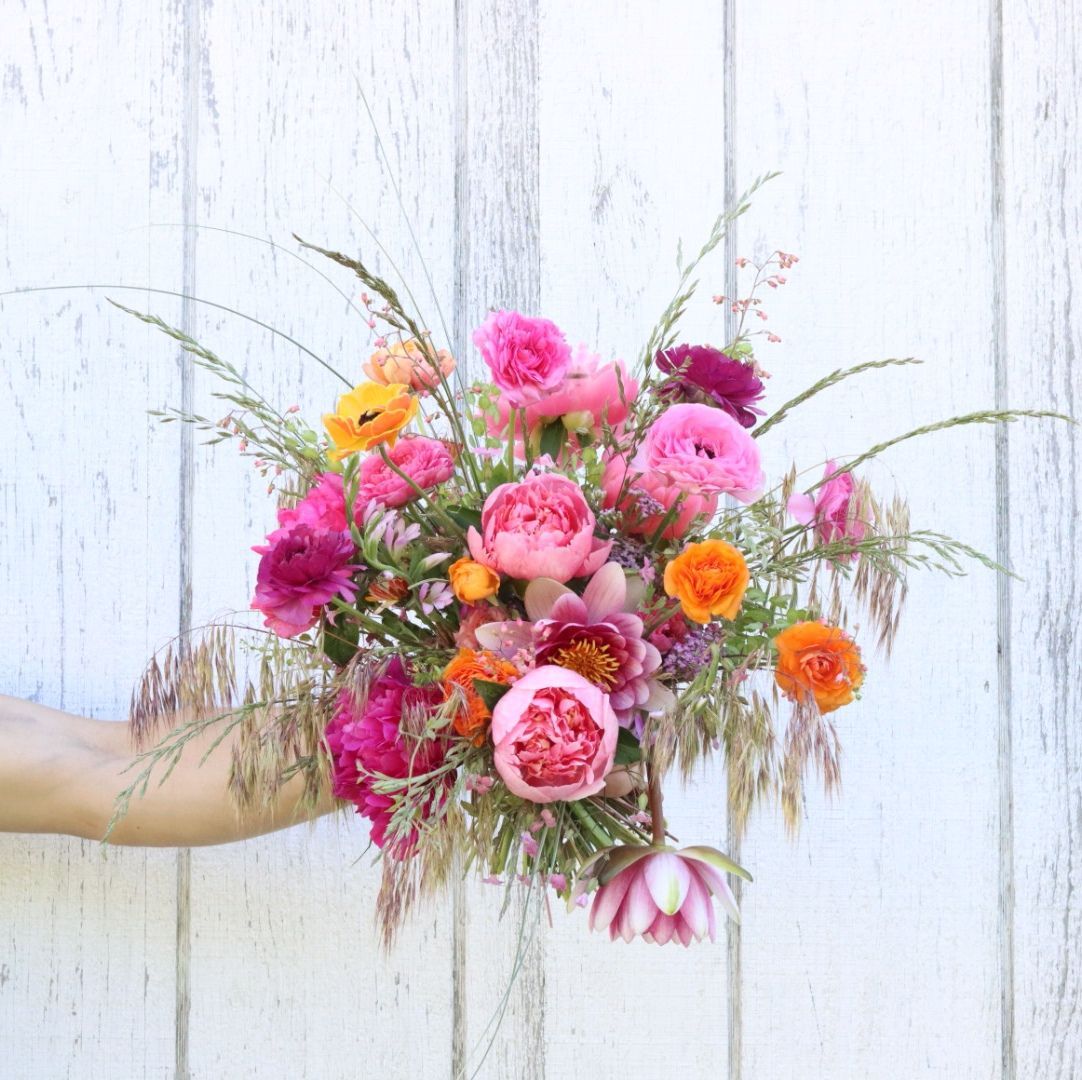 A person is holding a bouquet of pink and orange flowers in front of a white wooden wall.