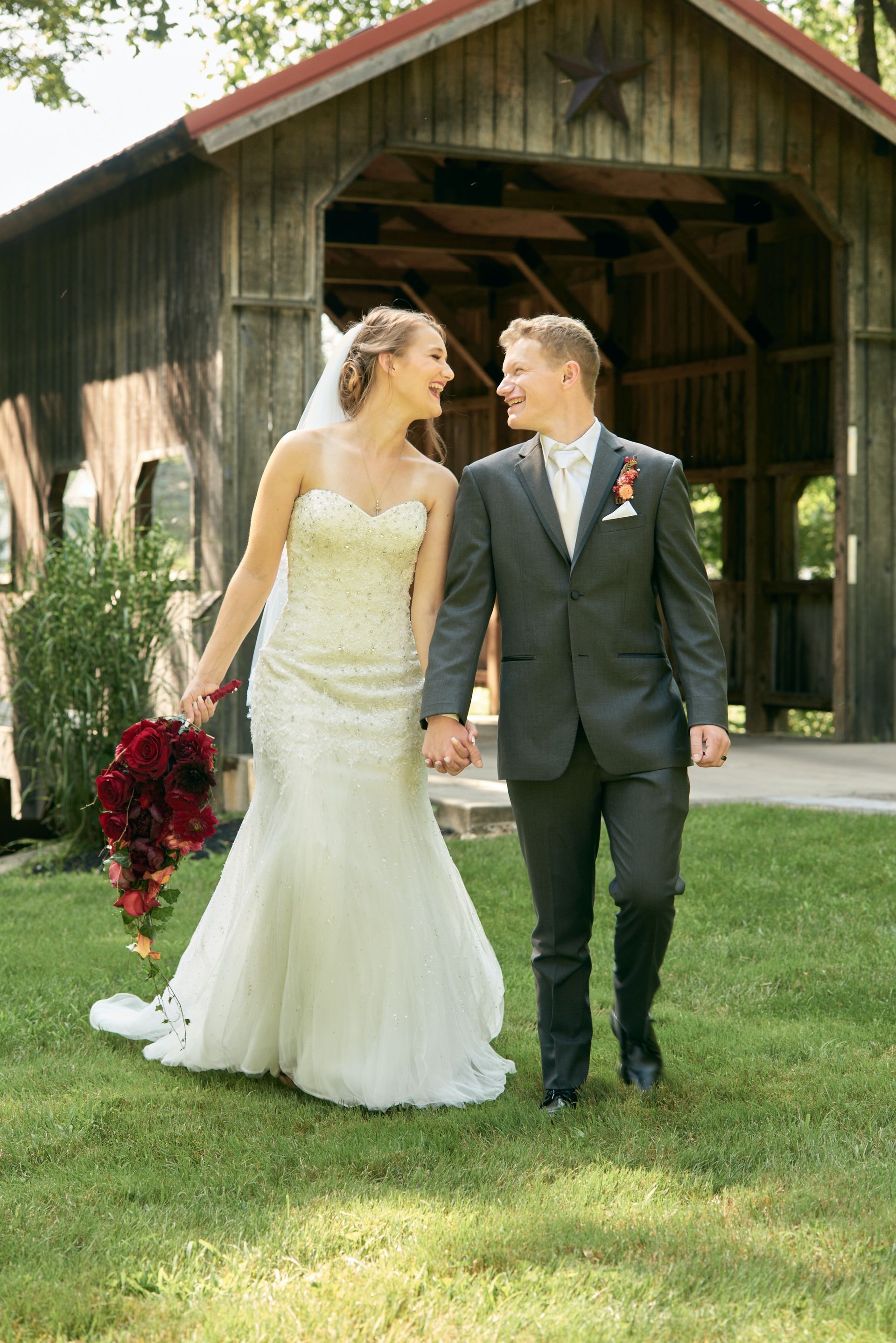 A bride and groom are holding hands and walking in front of a covered bridge.
