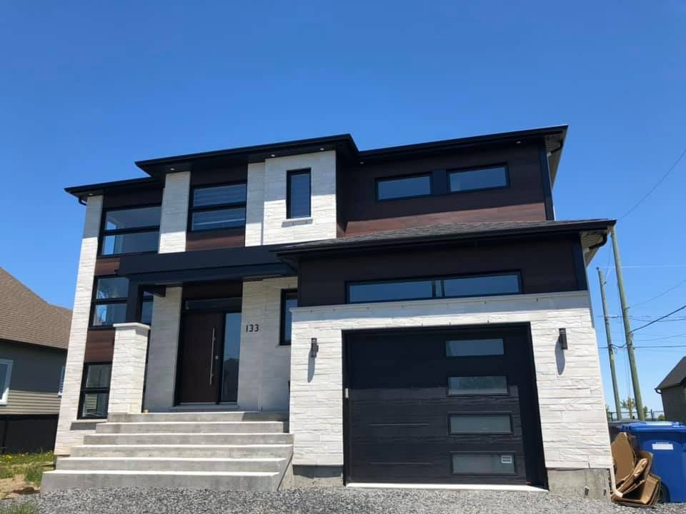 Une grande maison avec une porte de garage noire et une poubelle bleue devant.