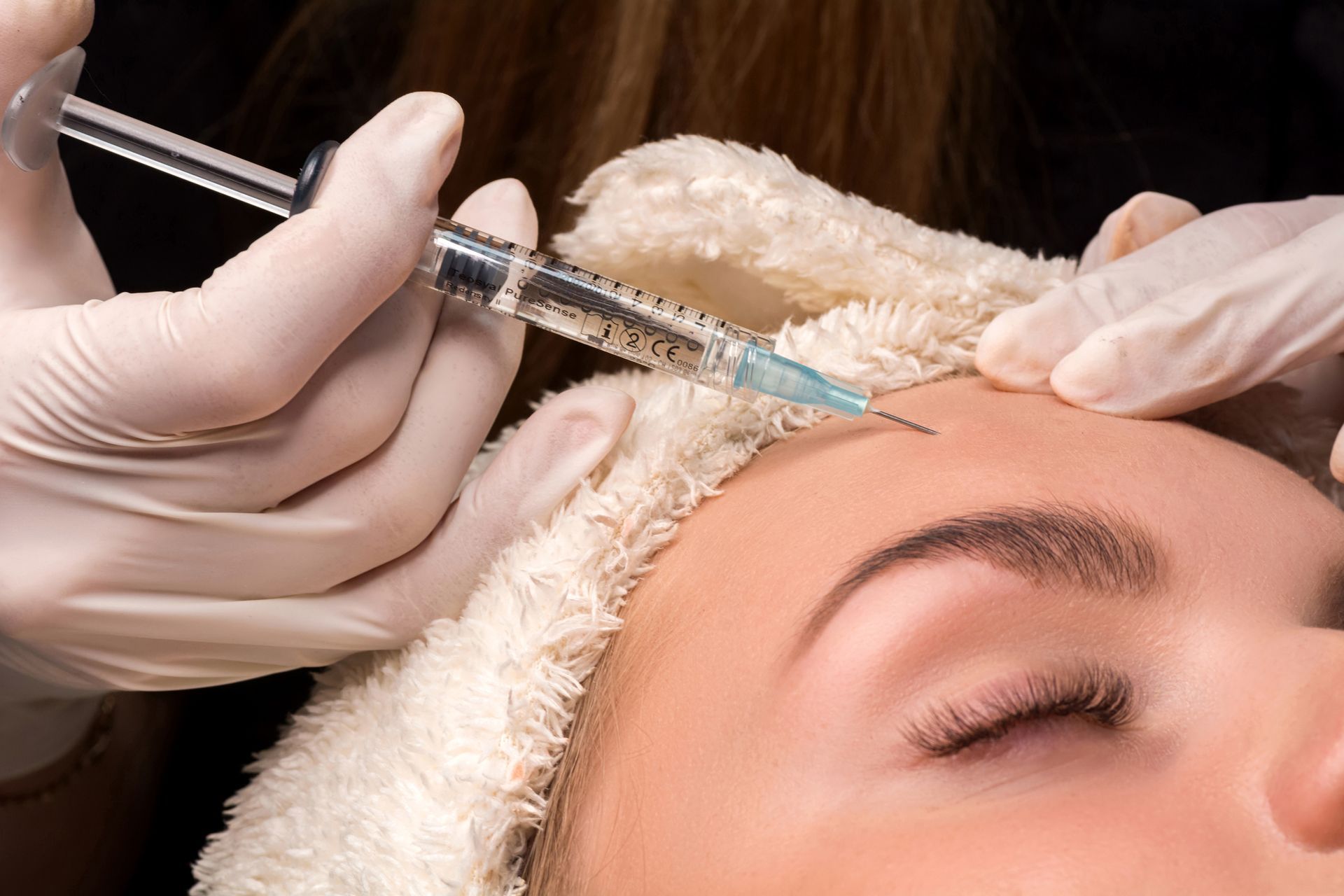 Person receiving cosmetic injections in the forehead; gloved hands, syringe, close-up.