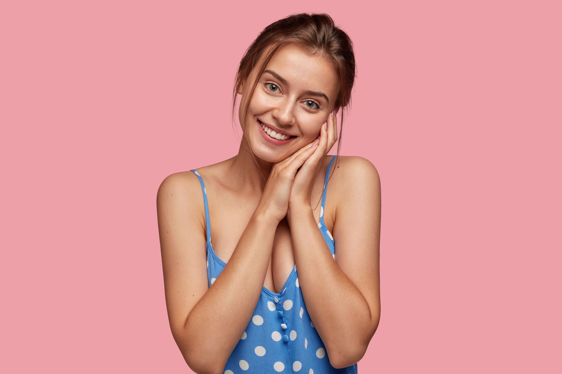 A woman in a blue polka dot dress is smiling with her hands on her face.