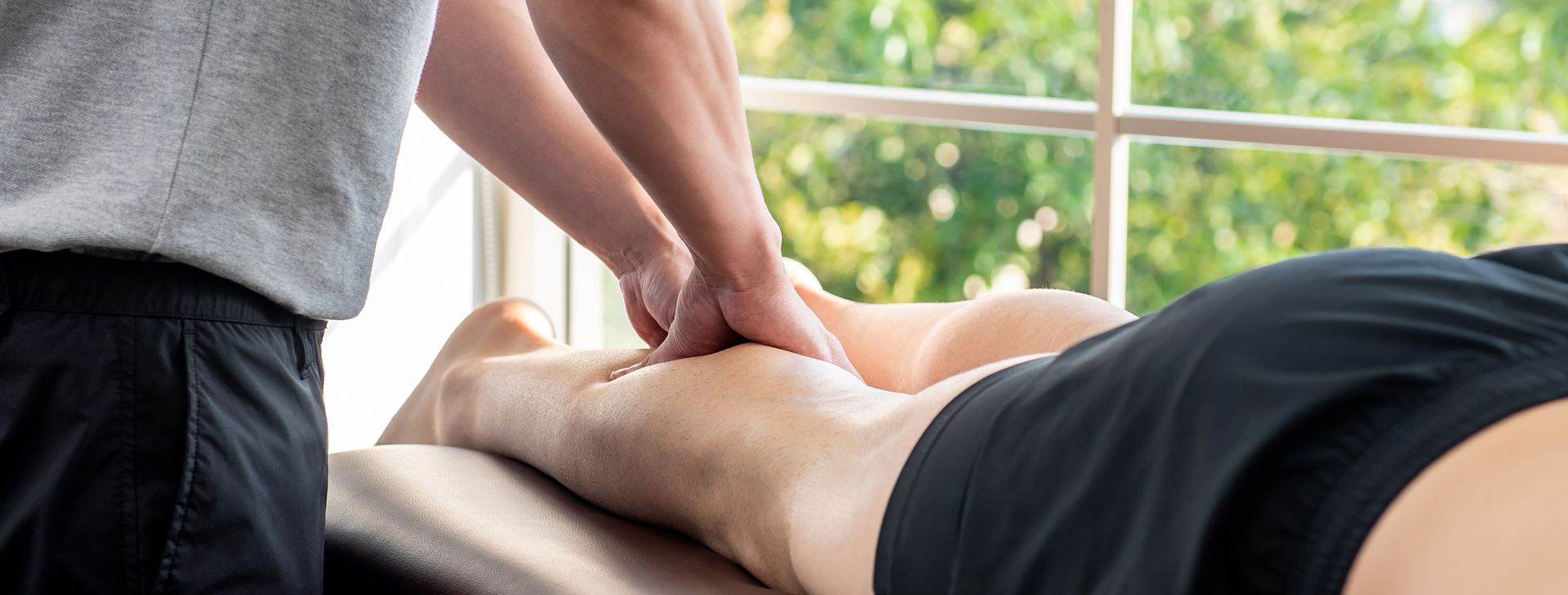 A person receiving a leg massage from a masseuse near a window with greenery.