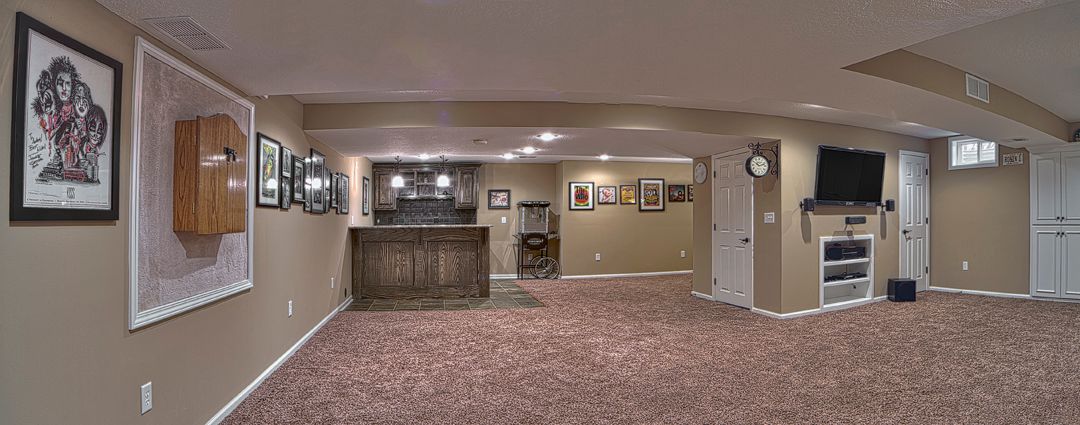 A panoramic view of a finished basement with beige walls, patterned carpeting, a wet bar, and a wall-mounted television.