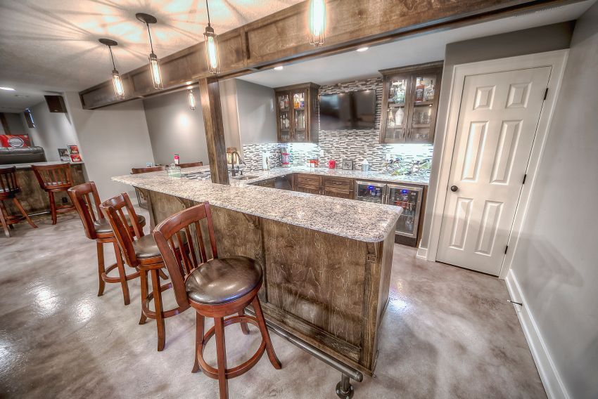A basement bar area with a granite countertop, rustic wooden cabinets, three bar stools, and a white paneled door.