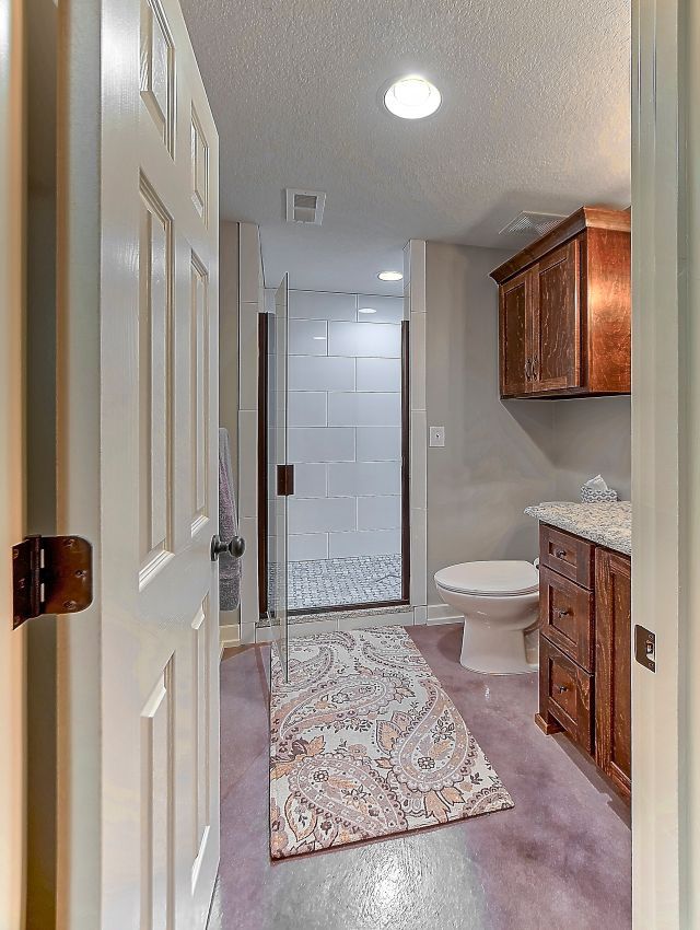 A bathroom with wood vanity cabinets, a white toilet, and a walk-in shower with white tiled walls and a paisley floor rug.