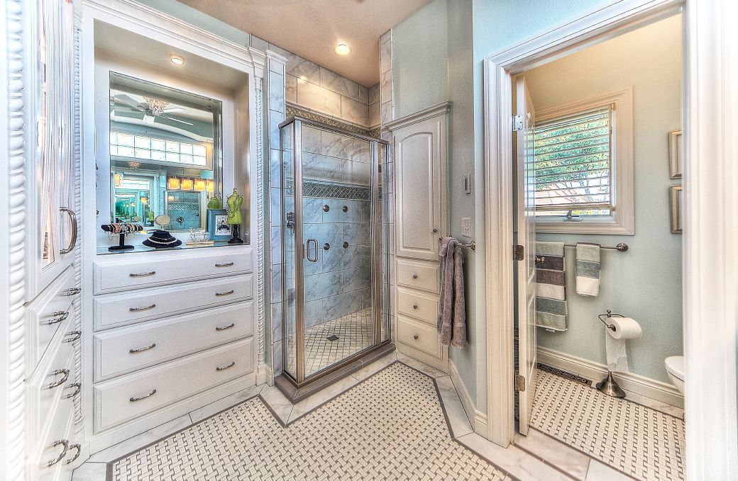 A bright, white bathroom with a built-in vanity, a glass-enclosed shower, patterned floor tiles, and an adjacent toilet.