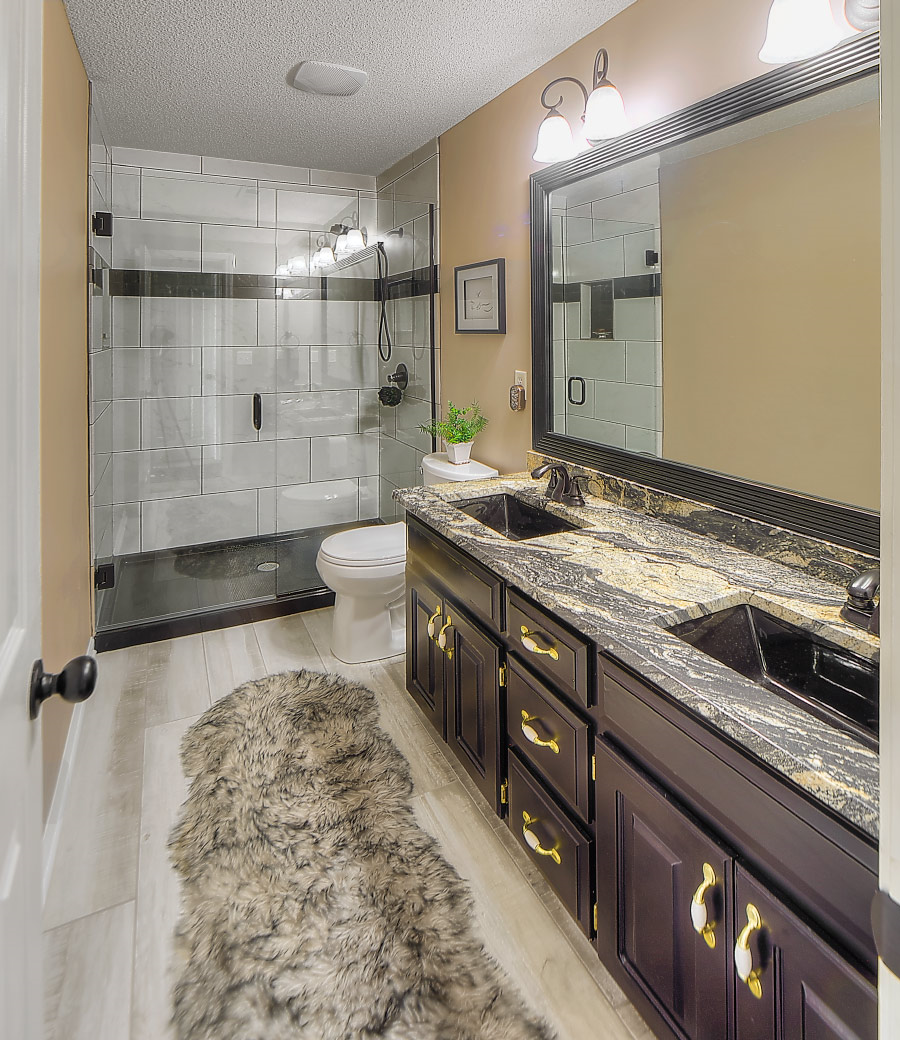 A modern bathroom with dark wood cabinets, granite countertops, a large mirror, a fuzzy rug, and a glass-enclosed shower.