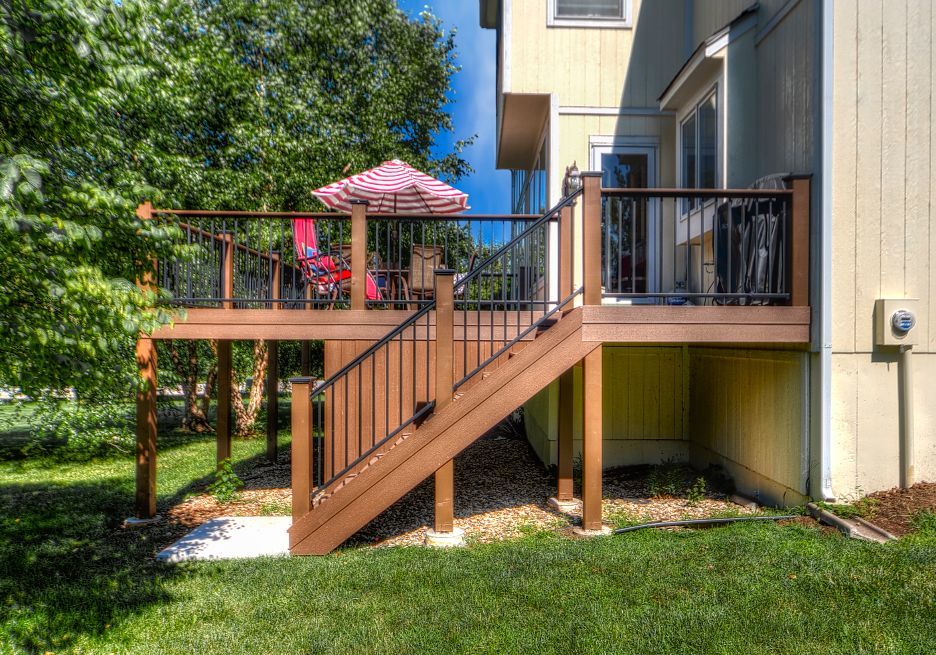A wooden deck with black railings and a striped patio umbrella, featuring stairs leading down to a grassy backyard.