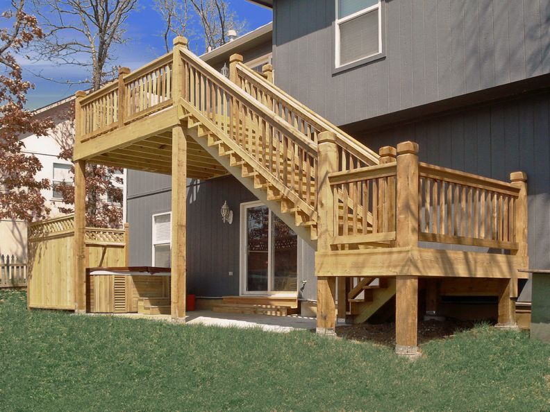 A large wooden deck with a staircase leads from an upper-level home entrance to a lower deck area with a hot tub.