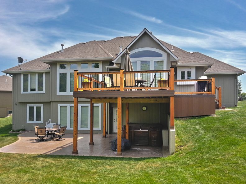 A two-story tan house with a large wooden deck overlooking a patio area and a grassy backyard under a blue sky.