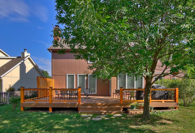 A two-story brown house with a large wooden deck and a green tree in the front yard on a sunny day.