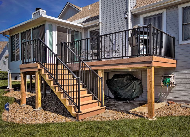 A wooden deck with black railings and stairs leads to a screened-in porch, with a covered grill stored underneath.