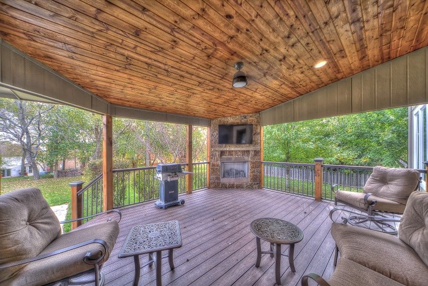 A covered outdoor deck with wood paneling, featuring a stone fireplace, television, grill, and lounge furniture.
