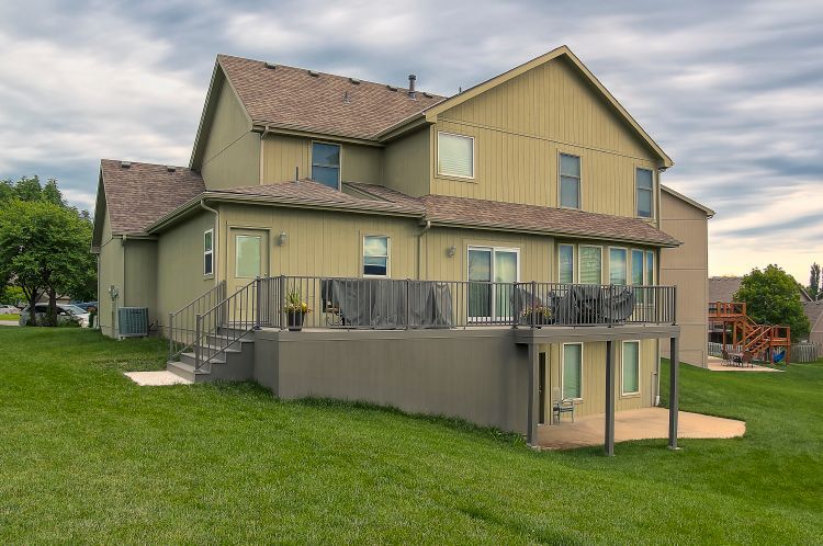 A two-story olive green house with a raised rear deck, gray structural supports, and an attached concrete patio below.