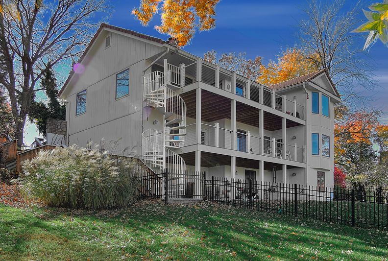 Three-story house with white siding and stacked back balconies, featuring a white spiral staircase against autumn trees.