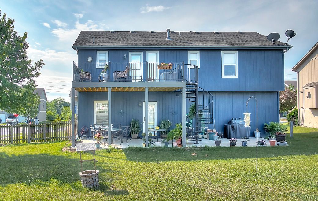 A two-story blue house with a second-floor deck, a spiral staircase, a patio area, and a large grassy backyard.