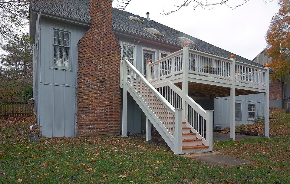 A gray house with a brick chimney and a large white wooden deck with stairs leading down to a lawn covered in autumn leaves.