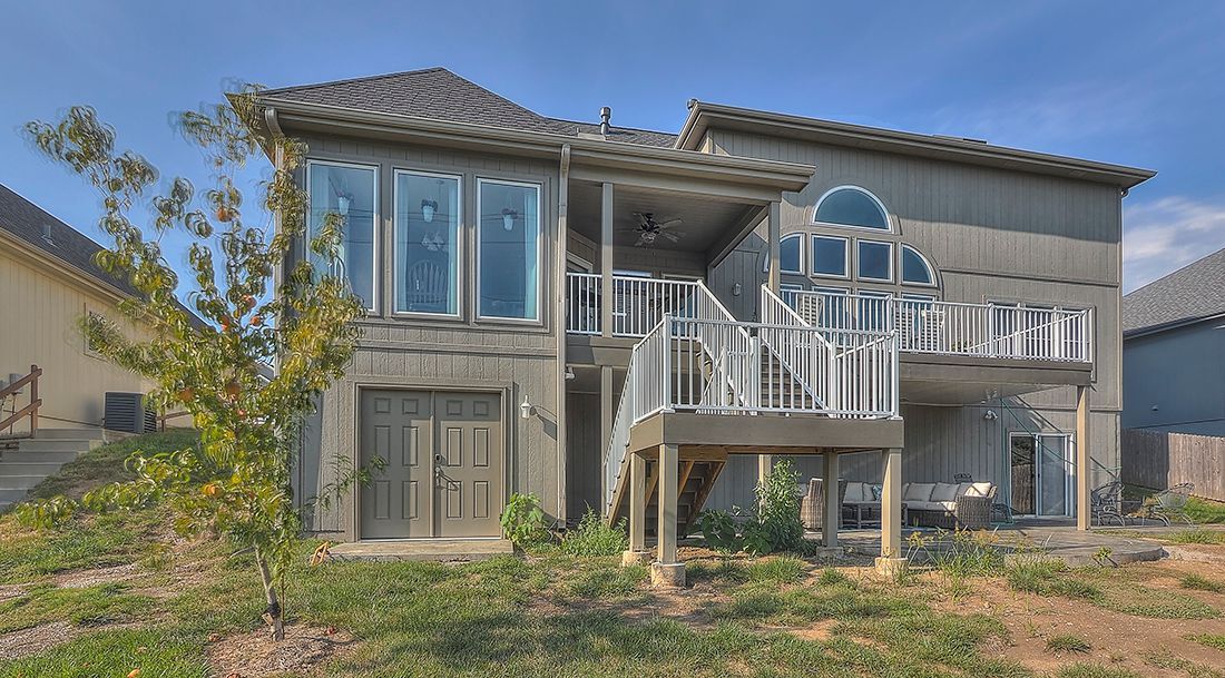 A two-story grey stucco house featuring an elevated deck with white railings, a lower patio, and a small tree in the yard.