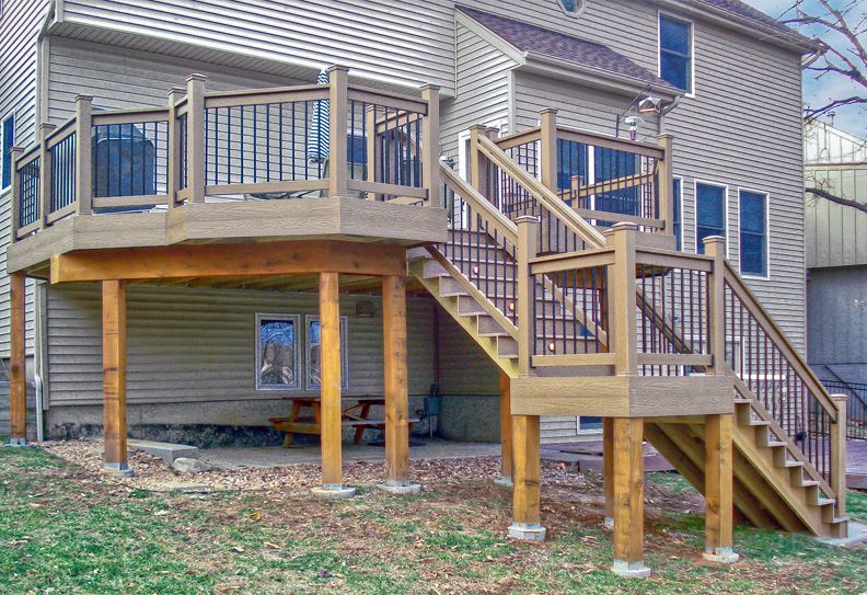 A raised wooden deck with a staircase and metal railings attached to the back of a beige two-story house.
