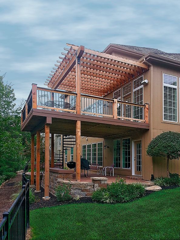 A two-story tan house features a large elevated wooden deck with a pergola, a spiral staircase, and a stone patio below.