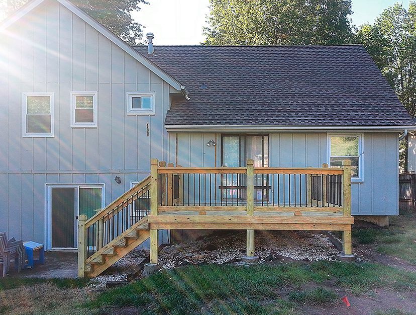 A light blue house with a newly constructed wooden deck featuring stairs and black metal railings in a grassy backyard.