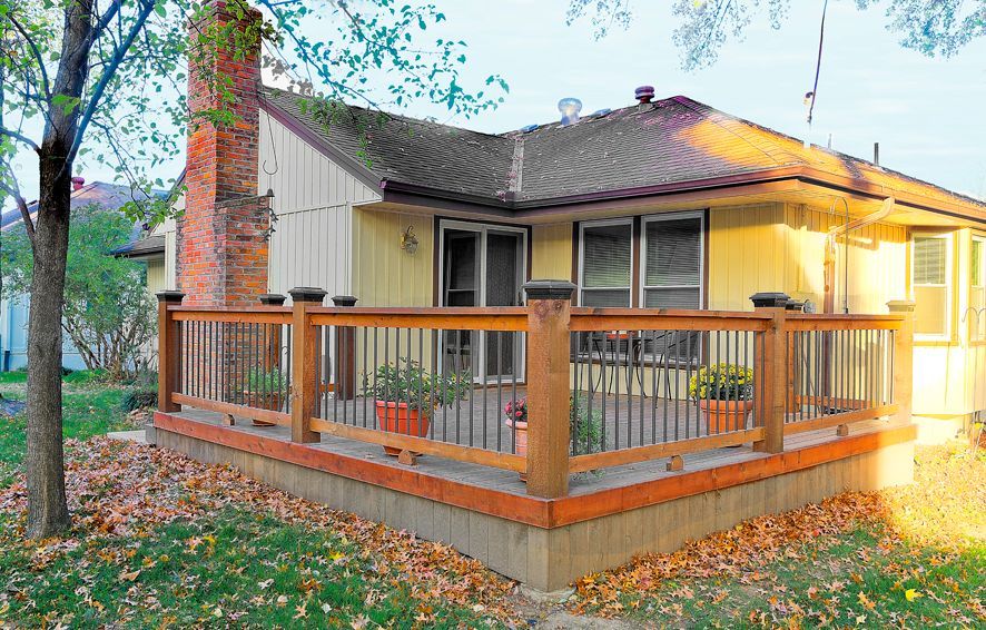 A light-yellow house with a wooden deck and brick chimney, surrounded by autumn leaves on a grassy yard.
