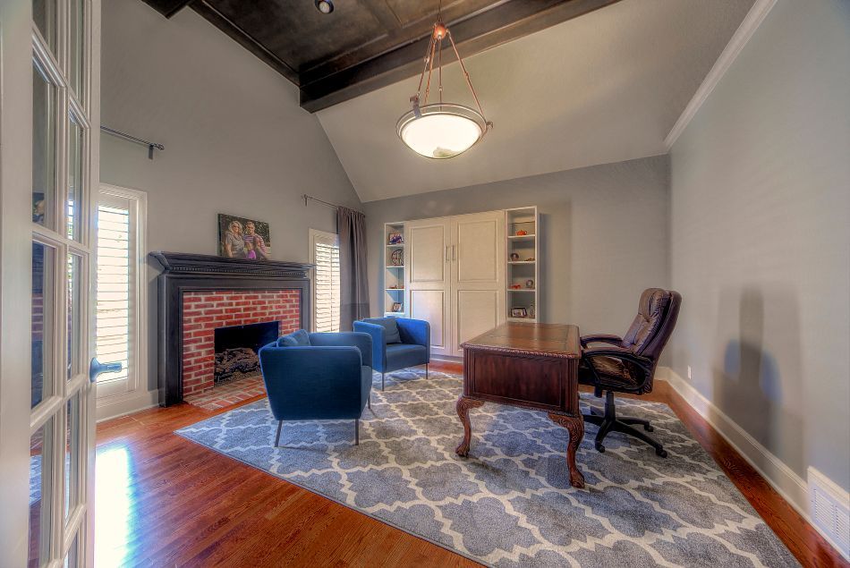 A home office with a brick fireplace, blue armchairs, a wooden desk with a rolling chair, and a patterned area rug.
