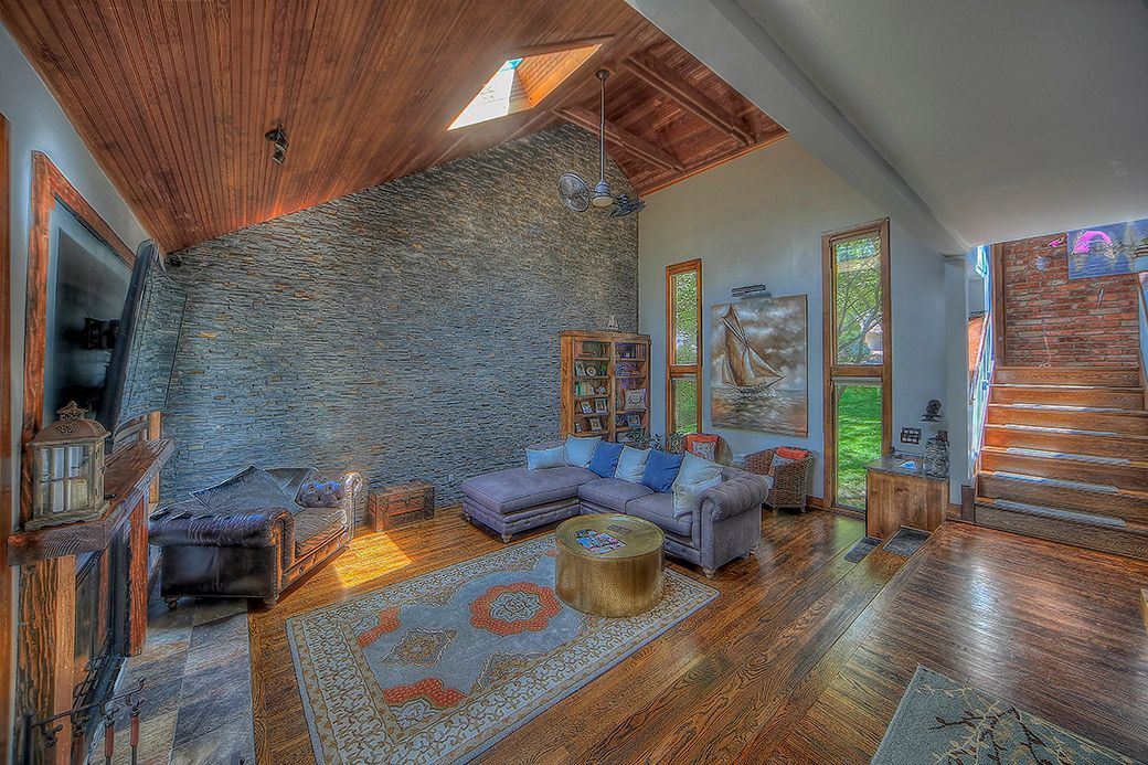A rustic living room with a large stone feature wall, vaulted wood ceiling, gray sectional sofa, and a patterned rug.