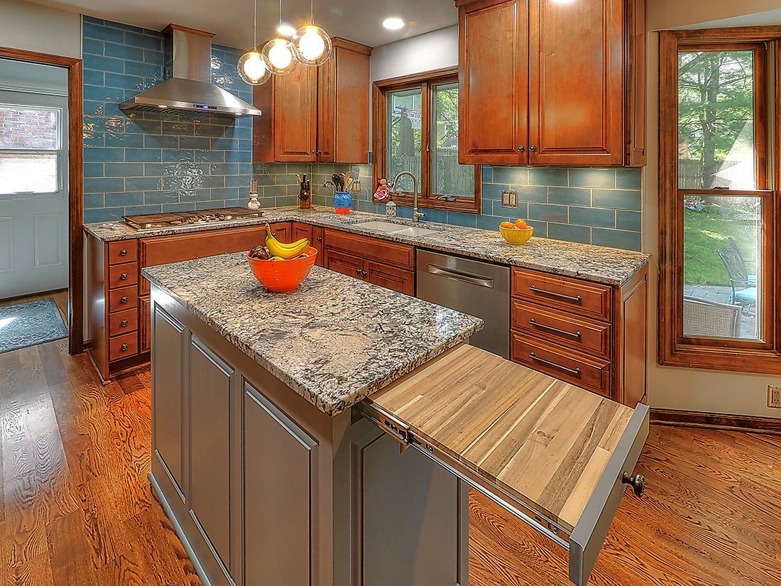 A rustic kitchen with wood cabinets, granite countertops, a grey island with a pull-out wooden board, and blue tile walls.