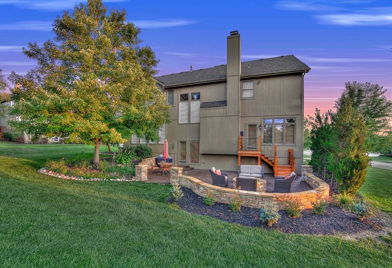 A beige two-story house with a wooden deck and a stone patio, surrounded by green grass and trees at sunset.