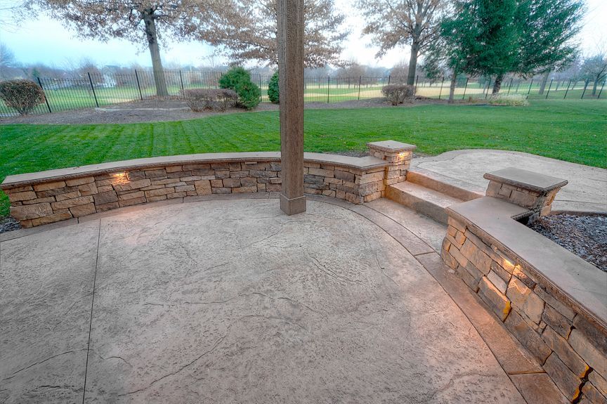 A curved stone patio wall with integrated lighting, overlooking a grassy backyard with trees under a cloudy sky.
