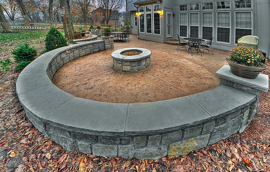 A curved stone seating wall encircles a central fire pit on a patio next to a house with large windows.