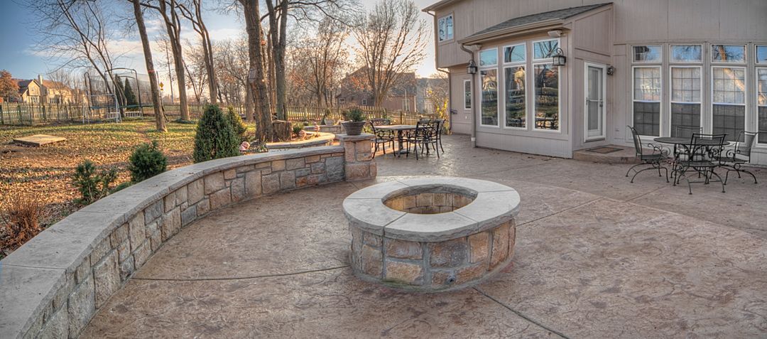 A stone fire pit sits on a stamped concrete patio outside a house with a pergola, stone retaining wall, and backyard.