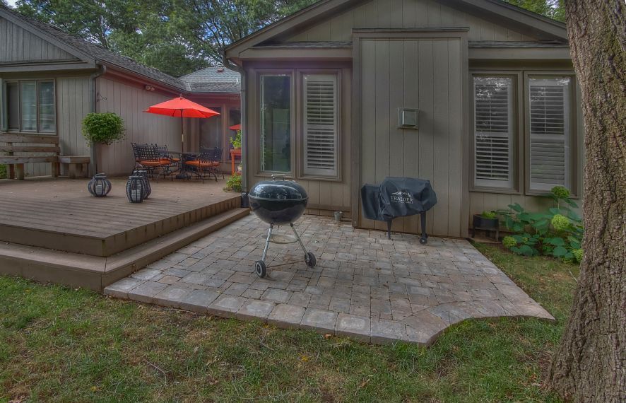 A stone patio with a charcoal grill beside a wooden deck and house with an orange umbrella in the background.