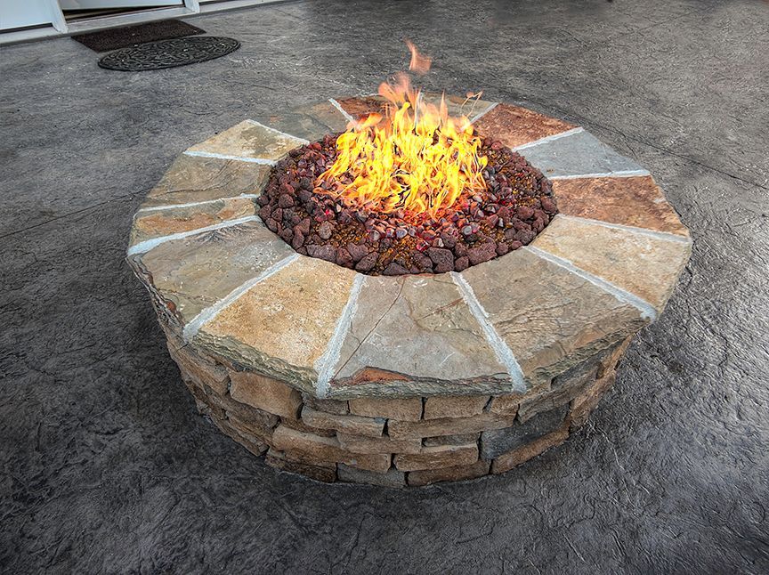 A circular stone fire pit with a blazing fire, placed on a dark, patterned patio.
