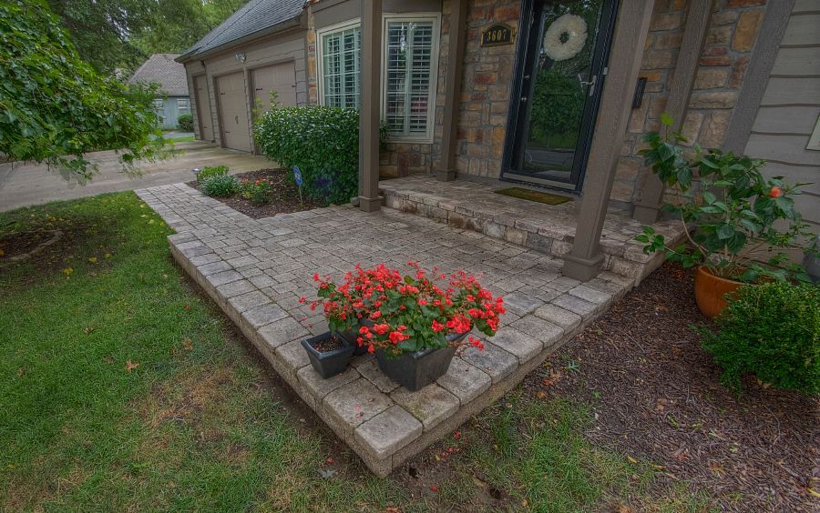 A stone patio with a container of orange flowers leads to a house entrance with stone siding and a dark front door.
