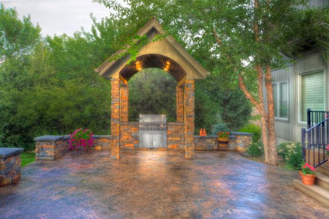 An outdoor stone patio featuring a covered grill area with a dark, patterned floor and lush green trees in the background.