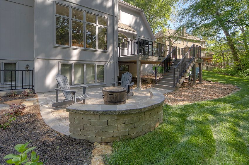 A beige stone patio with a built-in circular fire pit, two chairs, and a wooden deck with stairs leading to the lawn.