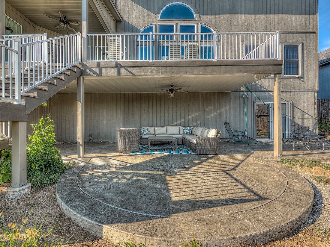 A modern house with a second-story deck over a shaded patio, featuring outdoor furniture on a circular concrete slab.
