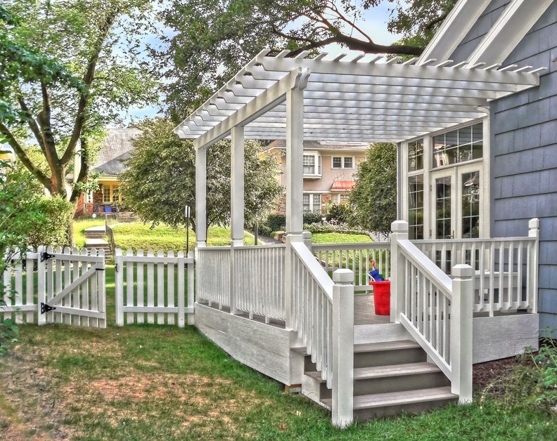 A raised white wooden deck with a matching pergola and picket fence attached to the side of a blue-sided house.
