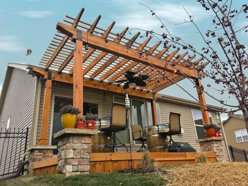 A wooden pergola covers a backyard deck featuring outdoor barrel seating against the back of a tan-sided house.