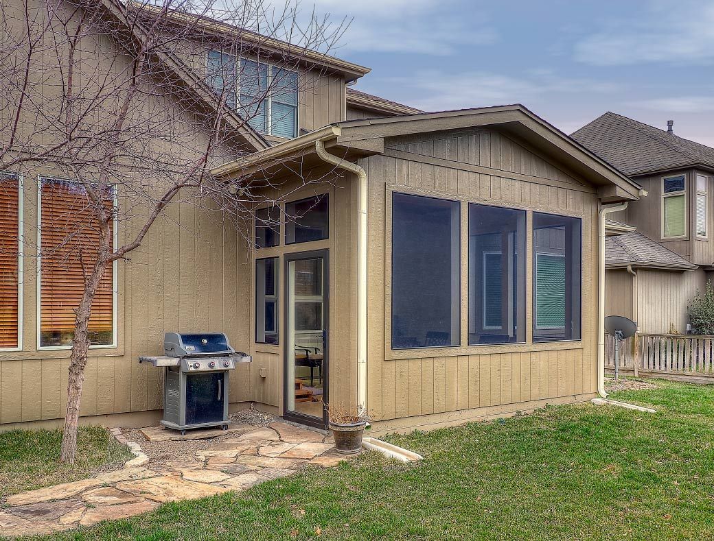 A beige house with a sunroom addition, a grill on the patio, and a grassy backyard.