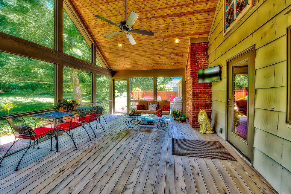 A sunroom with wood-paneled walls and ceilings, a brick chimney, large windows, patio furniture, and a ceiling fan.