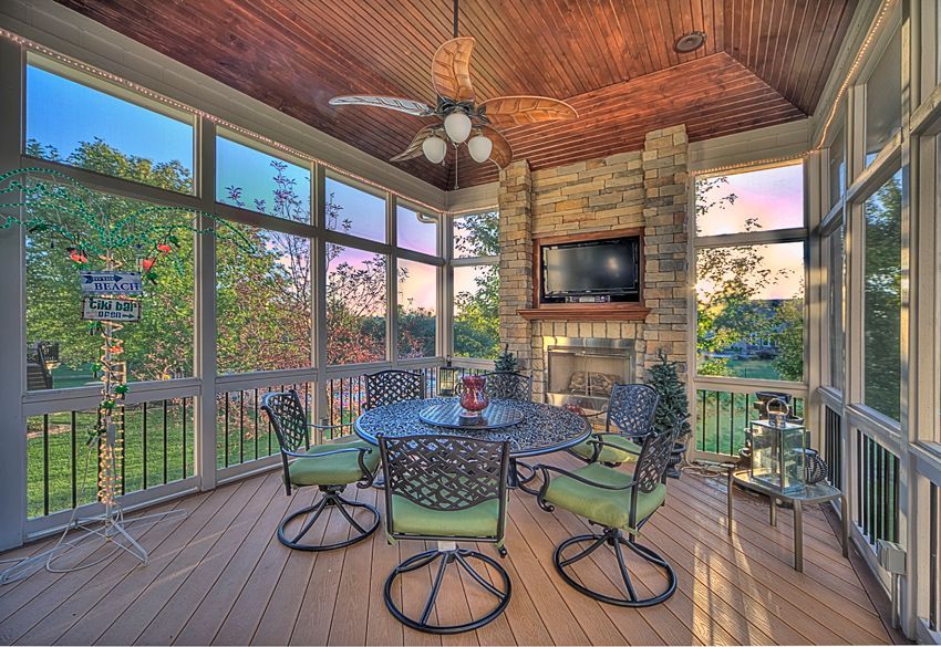 A screened-in porch with a wooden ceiling, stone fireplace, dining set, and sunset view through the windows.